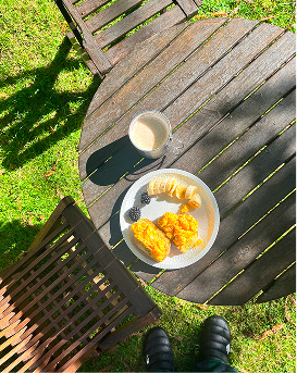 Desayuno al sol en una mesa de madera: café con leche, plátano, moras y dulces